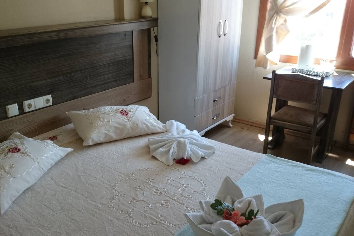 A neatly made bed with floral pillows and a decorative bow, a wooden headboard, a white wardrobe, and a wooden chair.