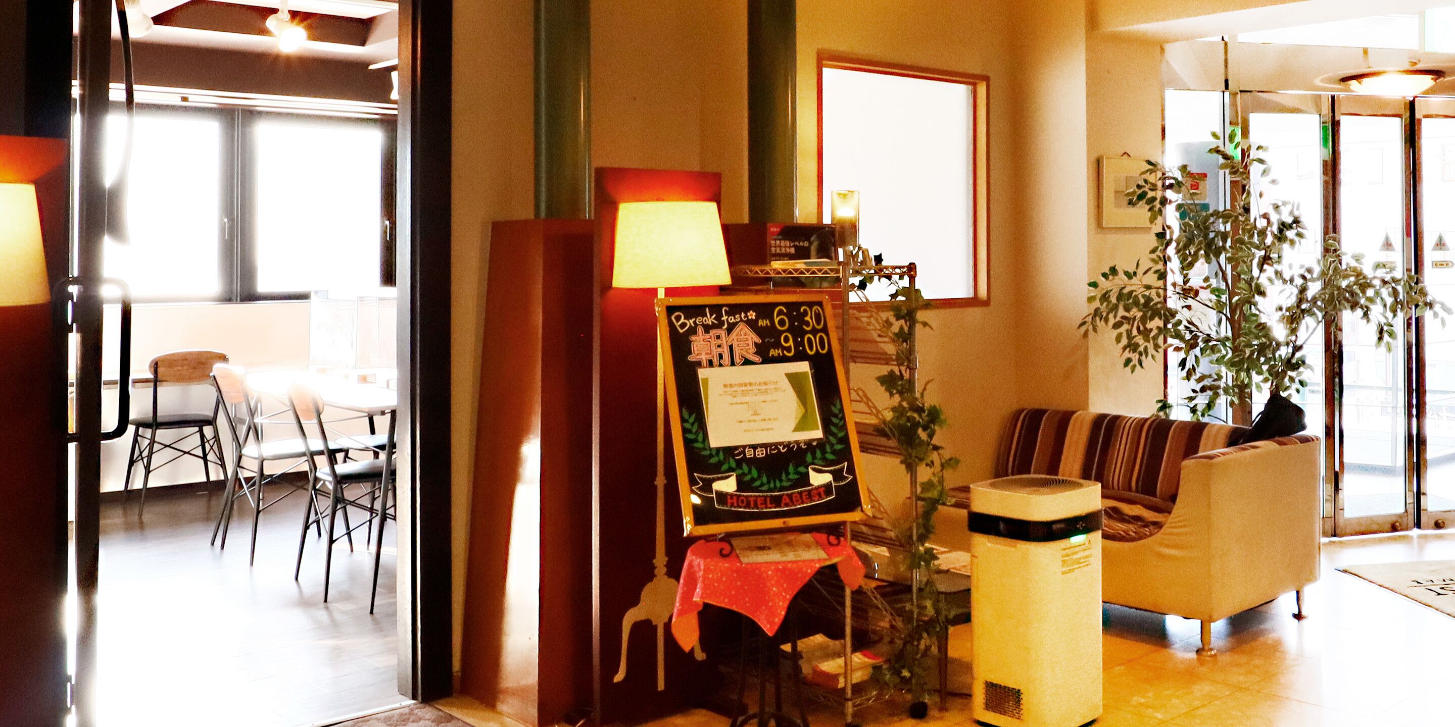 Vestíbulo de hotel con zona de comedor, un sofá, una mesa con lámpara y un cartel que indica los horarios del desayuno.