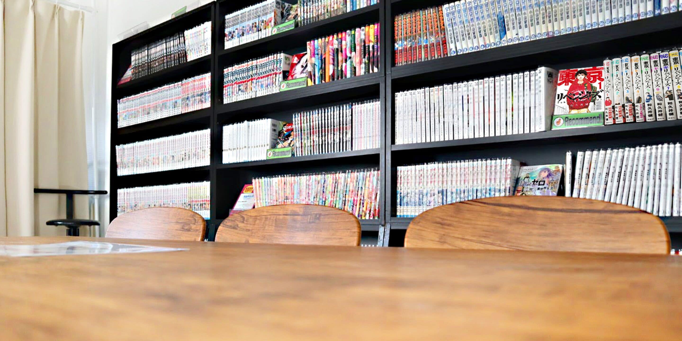 Une bibliothèque dont les étagères sont remplies de livres et de magazines. On aperçoit, au premier plan, une table entourée de chaises en bois.