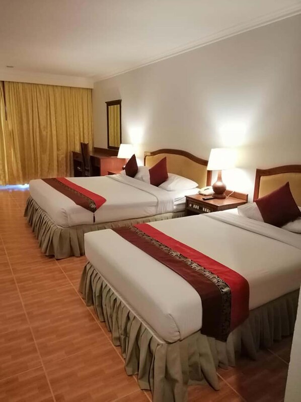 A hotel room with two beds, each with a wooden headboard, a bedside table with a lamp, and a curtain on the window.