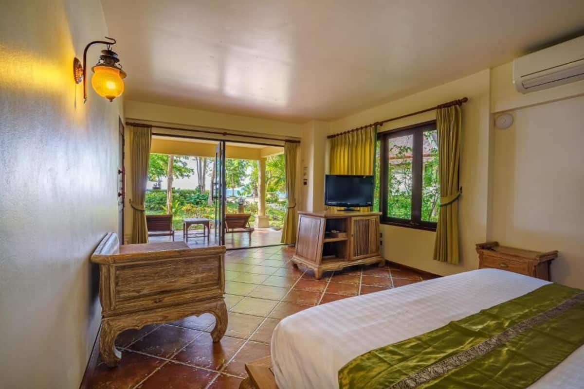 A hotel room with a bed, a TV, a wooden dresser, and a balcony with a view of greenery.