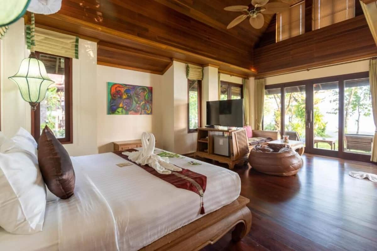 A hotel room with a wooden ceiling, a bed with white linens, a television, a desk with a computer, and a large window with a view of greenery.