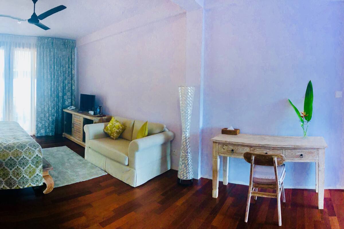 Uma sala de estar com sofá, mesa de centro, televisão, abajur, ventilador de teto, janela com cortinas e uma pequena mesa com uma planta.
