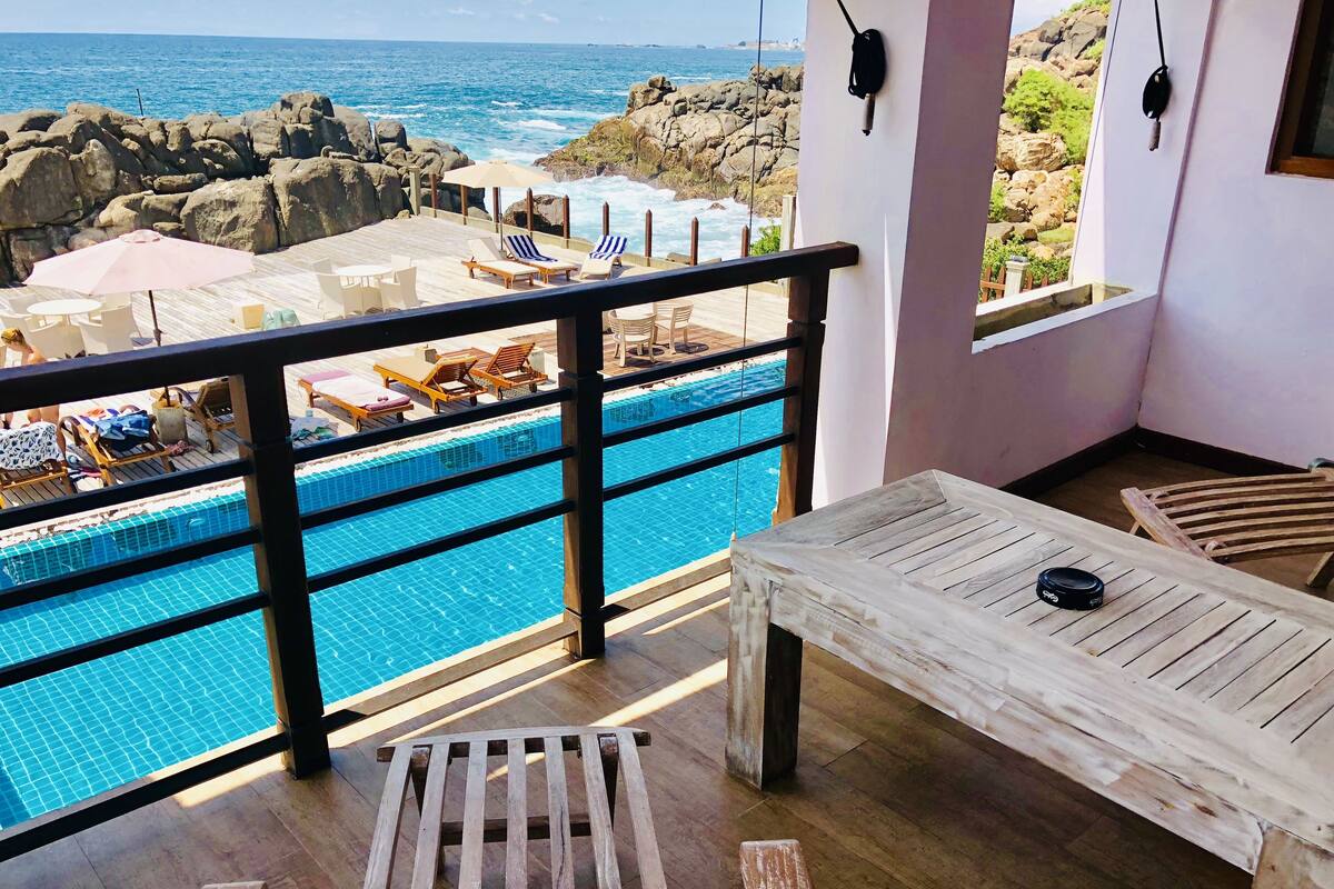 Varanda com vista para o oceano, uma piscina e mobiliário de madeira.