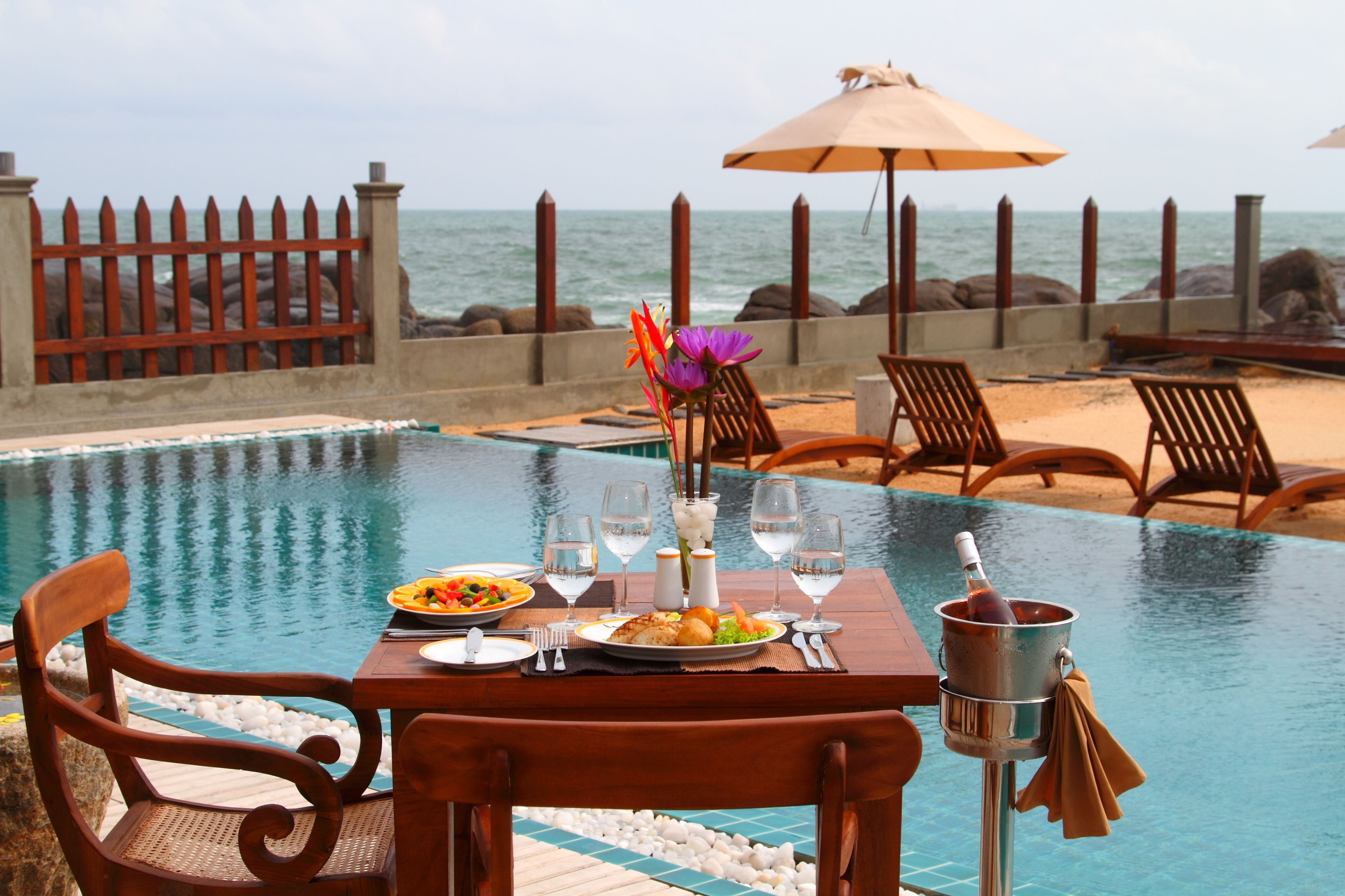 A dining table set with plates, glasses, and a bottle of champagne by a pool with a view of the ocean.
