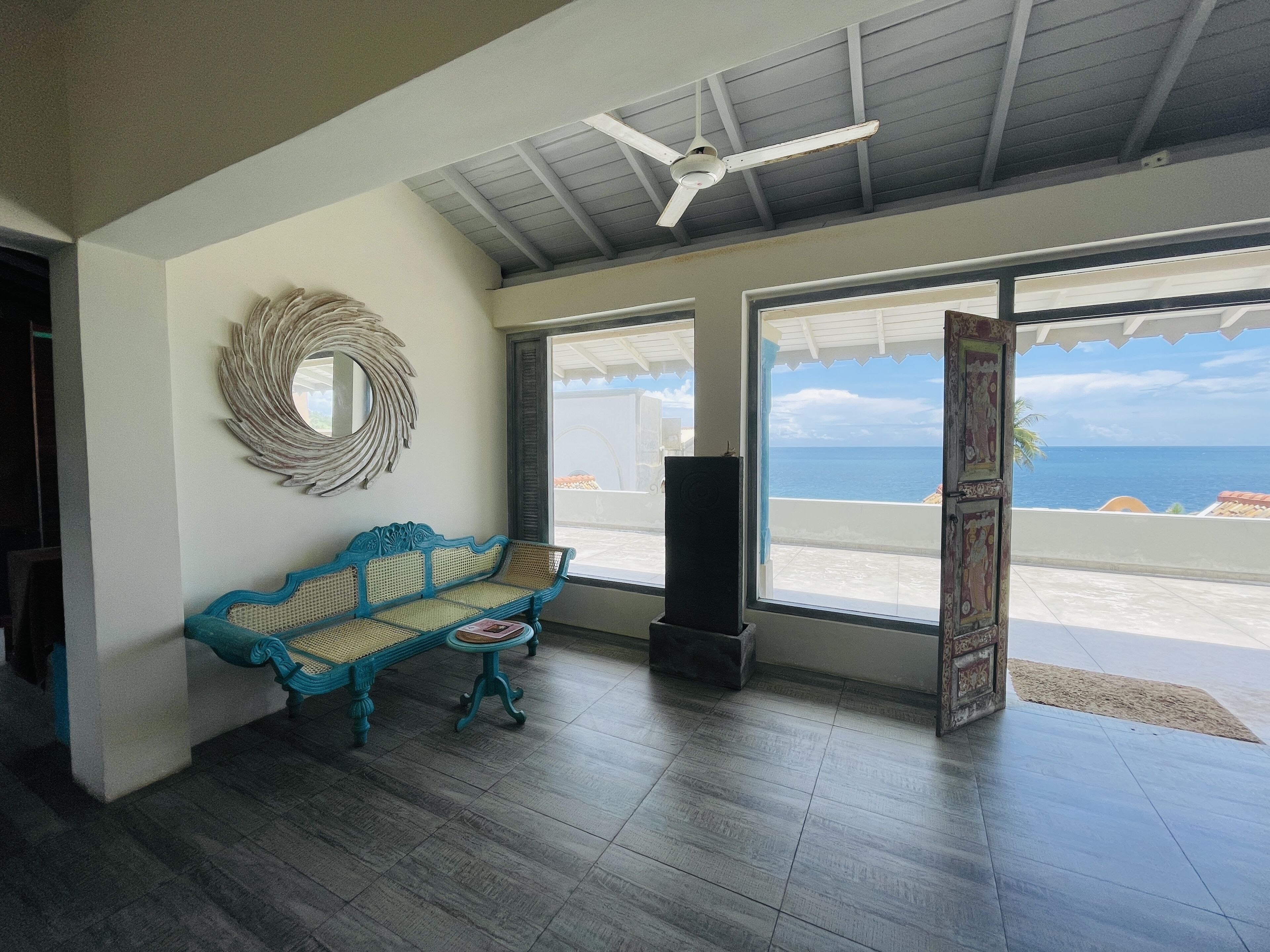 A room with a blue woven bench, a ceiling fan, and large windows offering a view of the ocean.