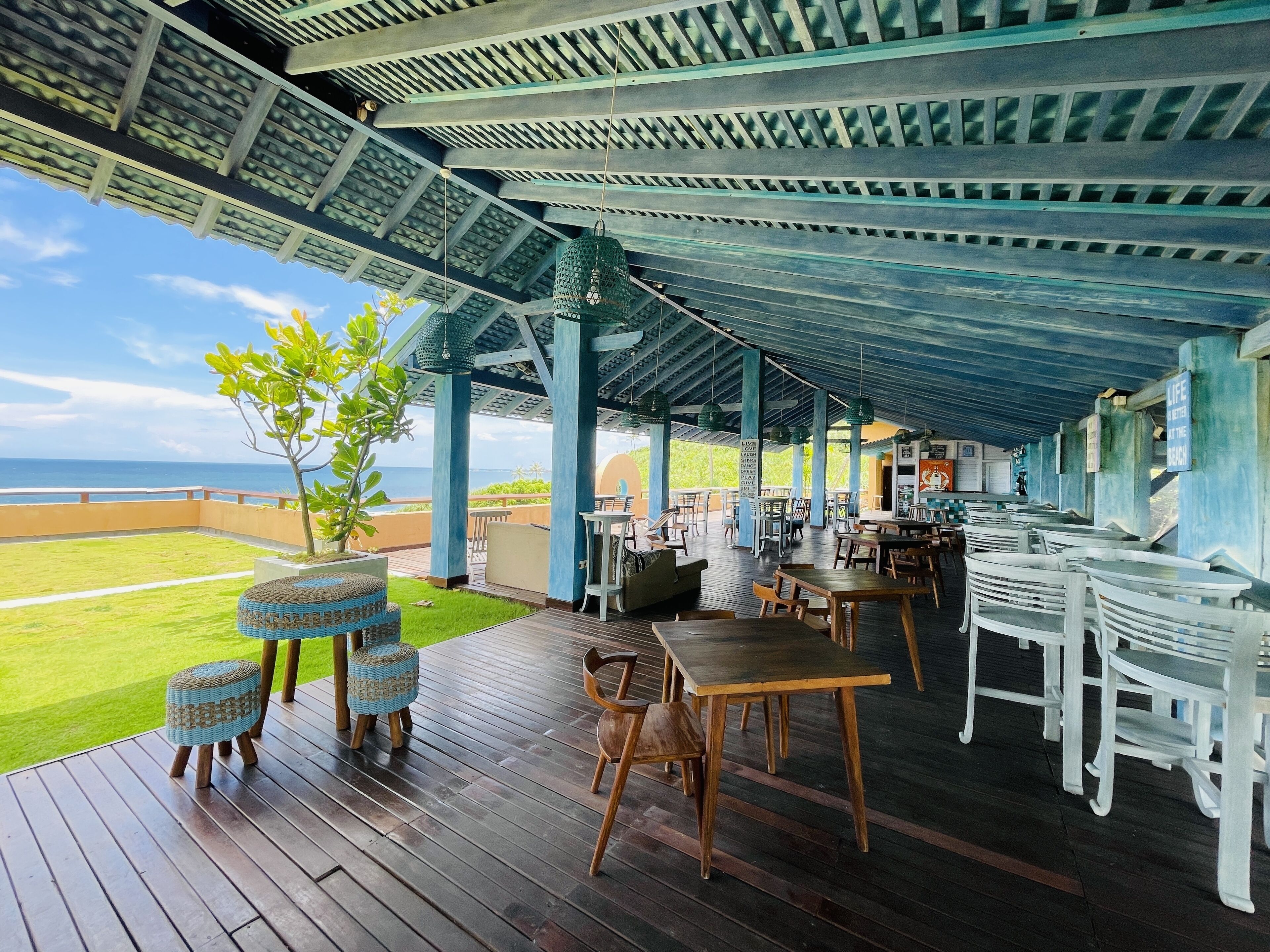 An outdoor seating area with wooden tables and chairs, a grassy lawn, and a view of the ocean.