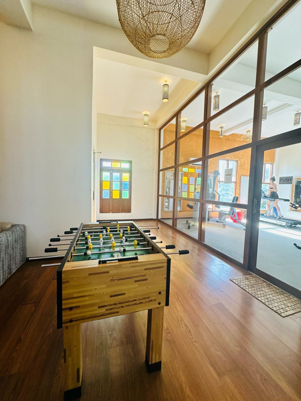 A room with a foosball table, large windows, and a view of a gym area.