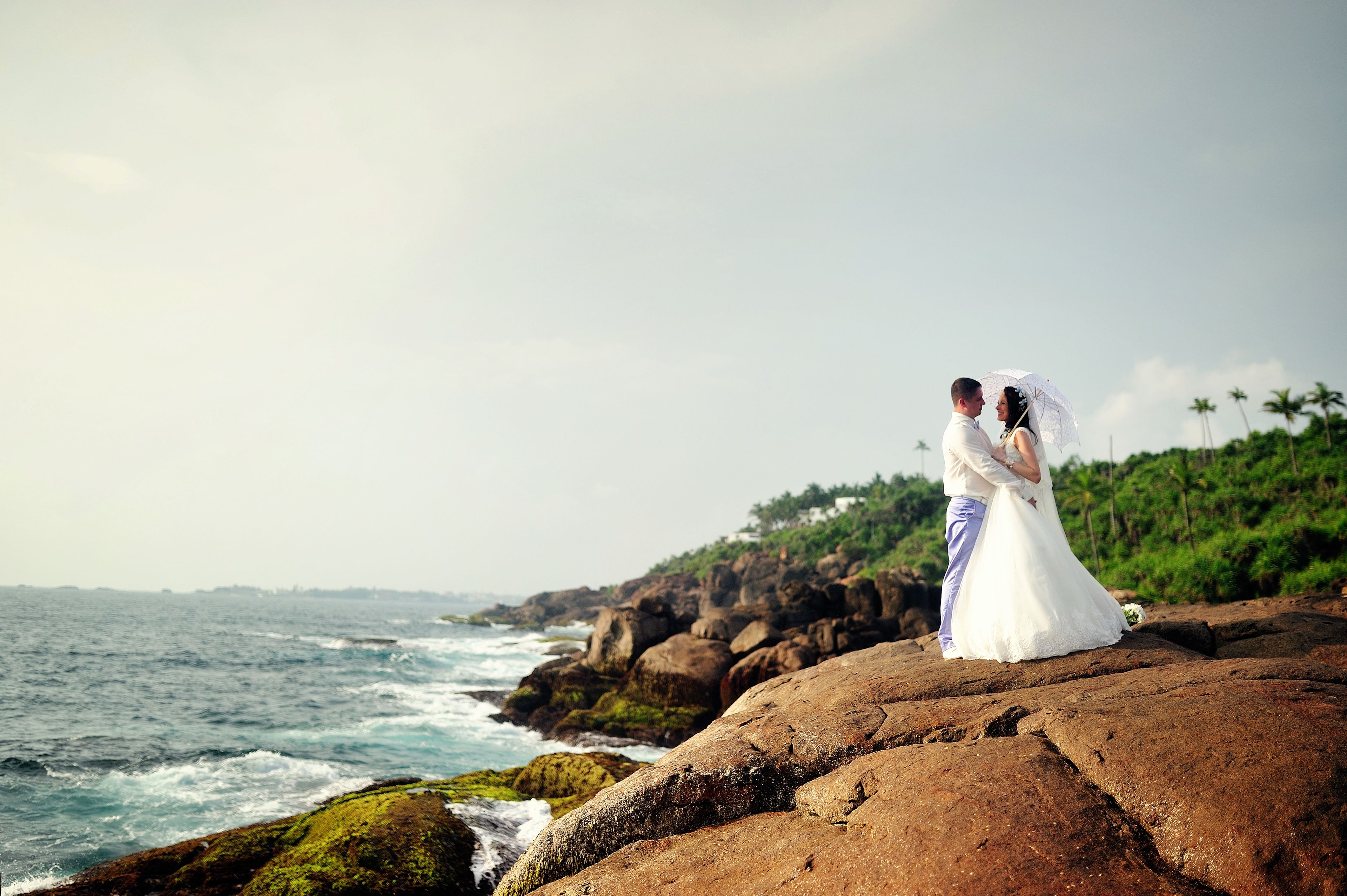A couple standing on a rocky outcrop by the sea with a view of the ocean and greenery in the background.