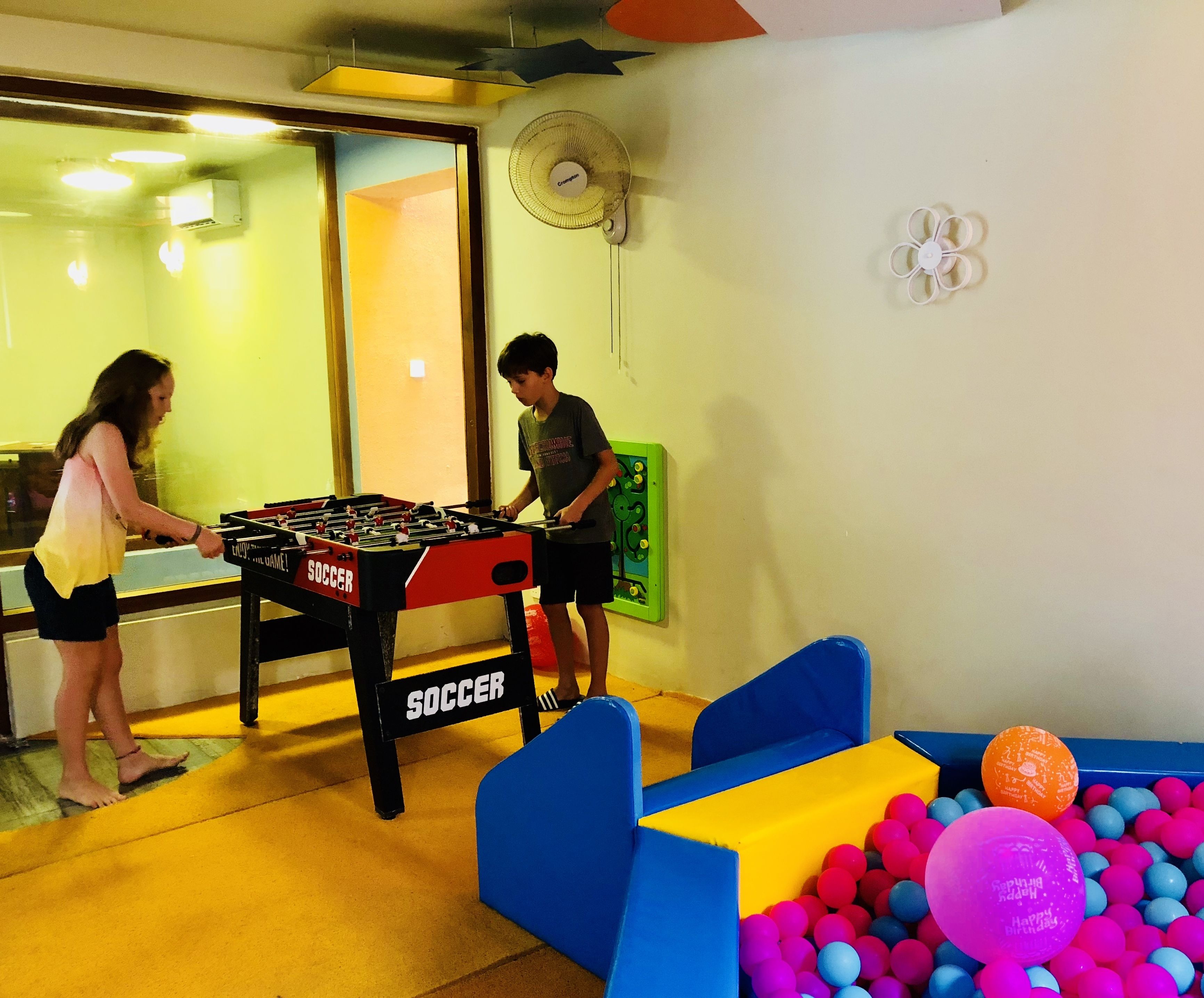 A room with a foosball table labeled "SOCCER," a ball pit with colorful balls, and a fan on the wall.