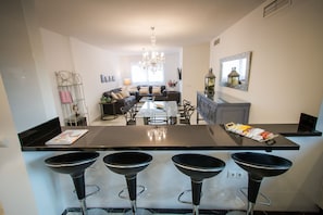 A modern kitchen with a central island, bar stools, and a dining area with a sofa and chandelier.