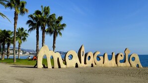 Ein Strand mit einer großen Skulptur, die "Malaga" aus beigem Material buchstabiert, Palmen und einem klaren, blauen Himmel.