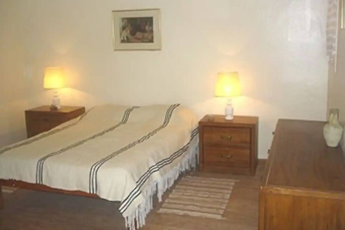 A room with a bed, two bedside tables with lamps, a small table with a vase, and a framed picture on the wall.