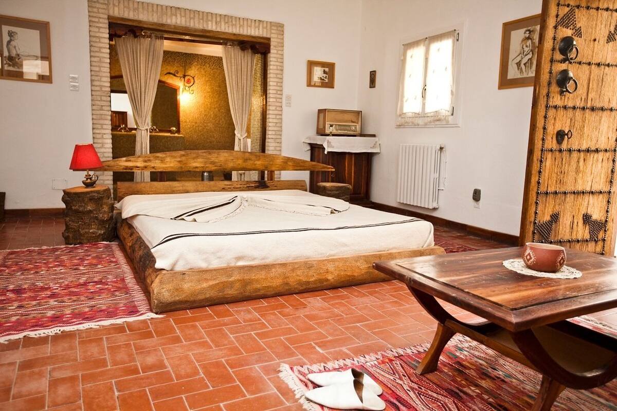 A bedroom with a wooden bed, a small table with a cup, a red lamp, and a patterned rug.