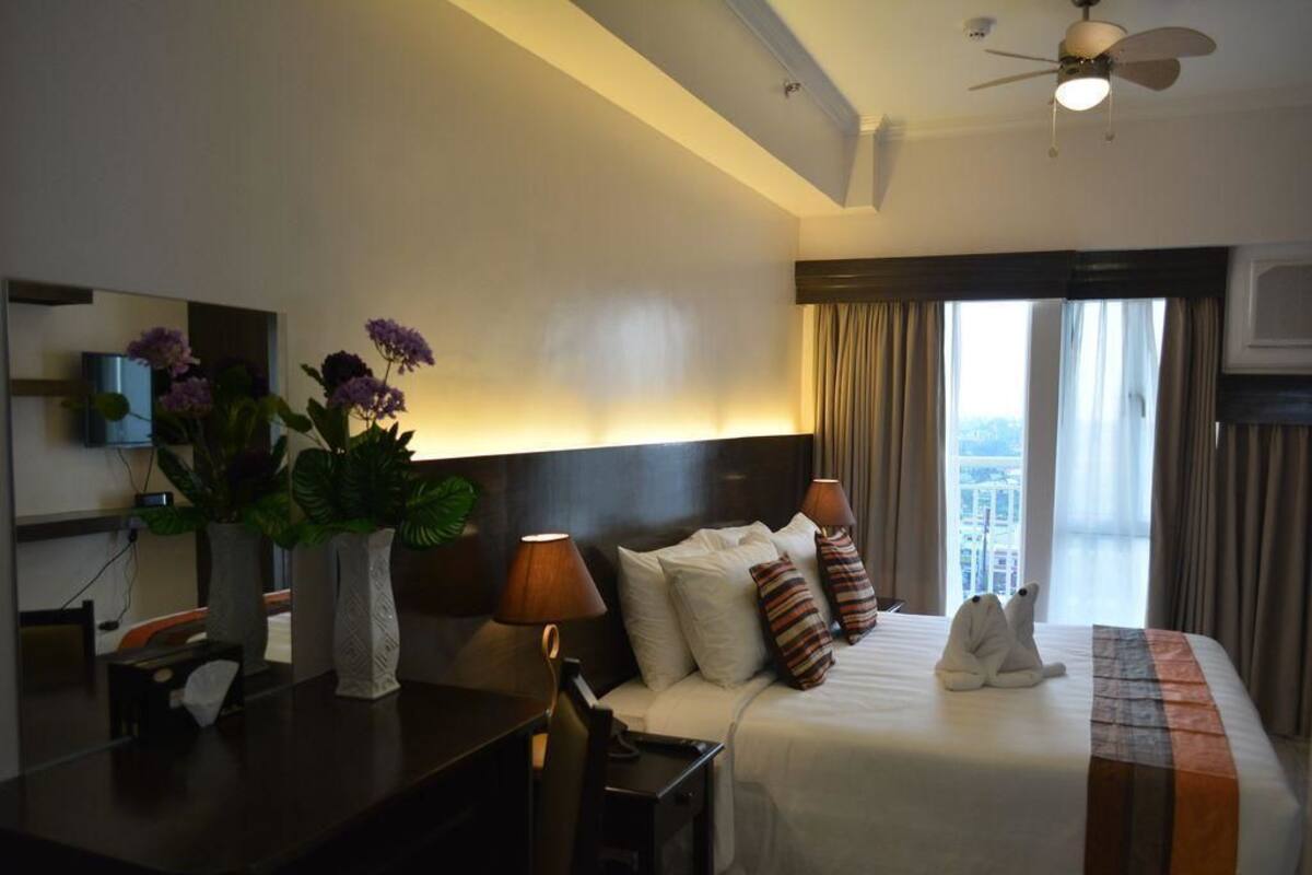 A hotel room with a bed, bedside table, lamp, and a view of the city through the window.