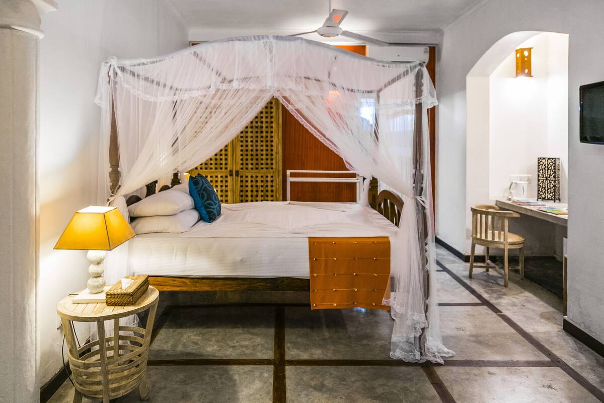 A room with a bed under a canopy, a bedside table with a lamp, a chair, and a desk with a chair.