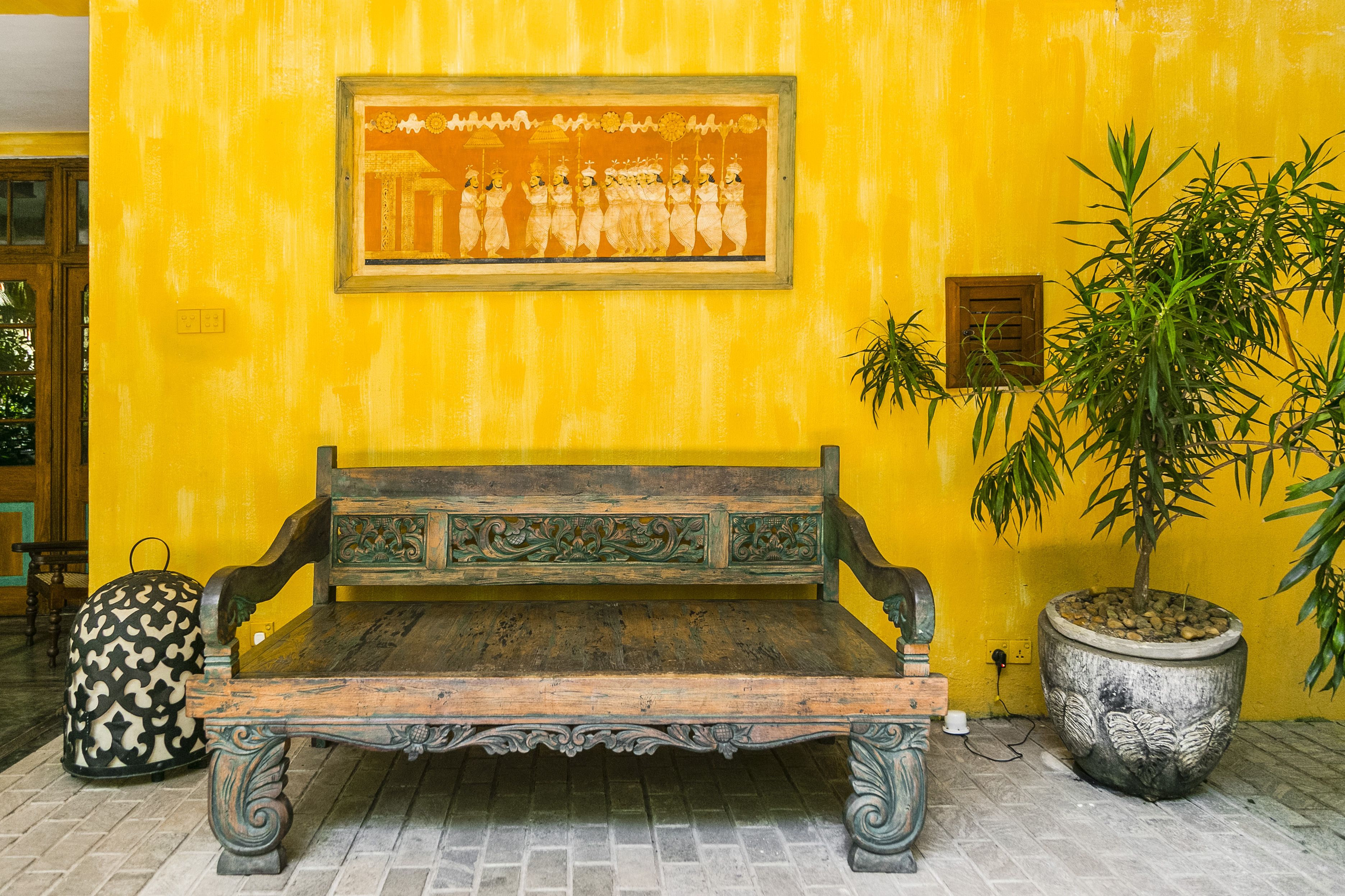 A wooden bench with intricate carvings, a large potted plant, and a framed artwork on a yellow wall.