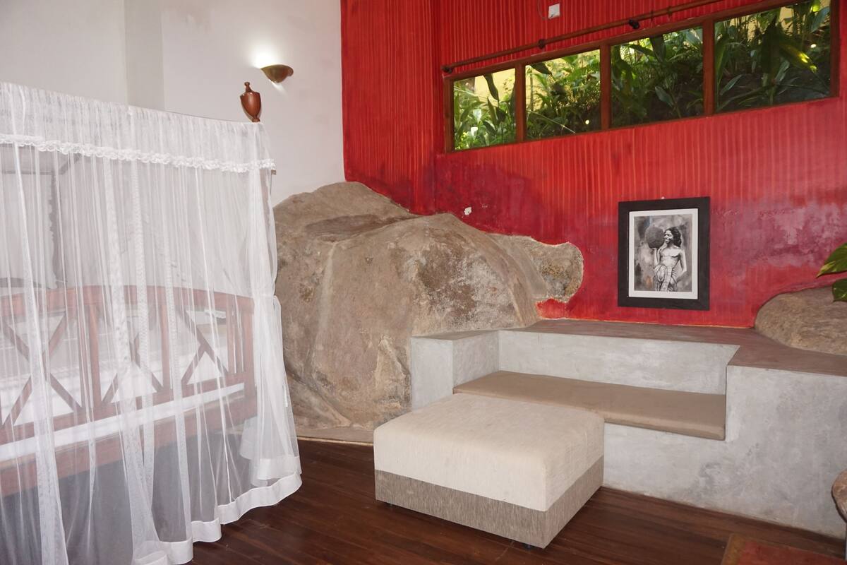 A room with a red wall, a framed picture, a large rock, and a white bench.
