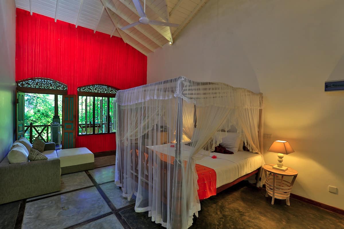 A room with a bed, a sofa, a table, and a window with a view of greenery.