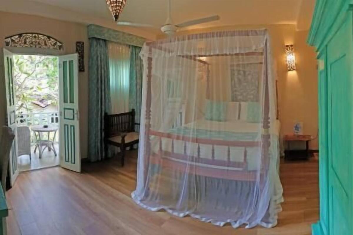 Quarto com cama, ventilador de teto, janela com cortinas e vista para o exterior.