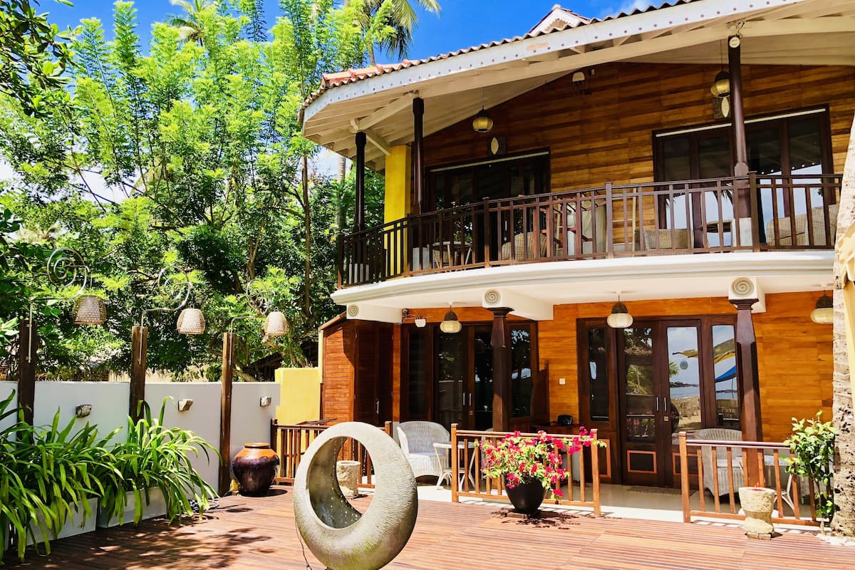 Uma casa de madeira de dois andares com varanda, rodeada por vegetação exuberante e plantas em vasos.