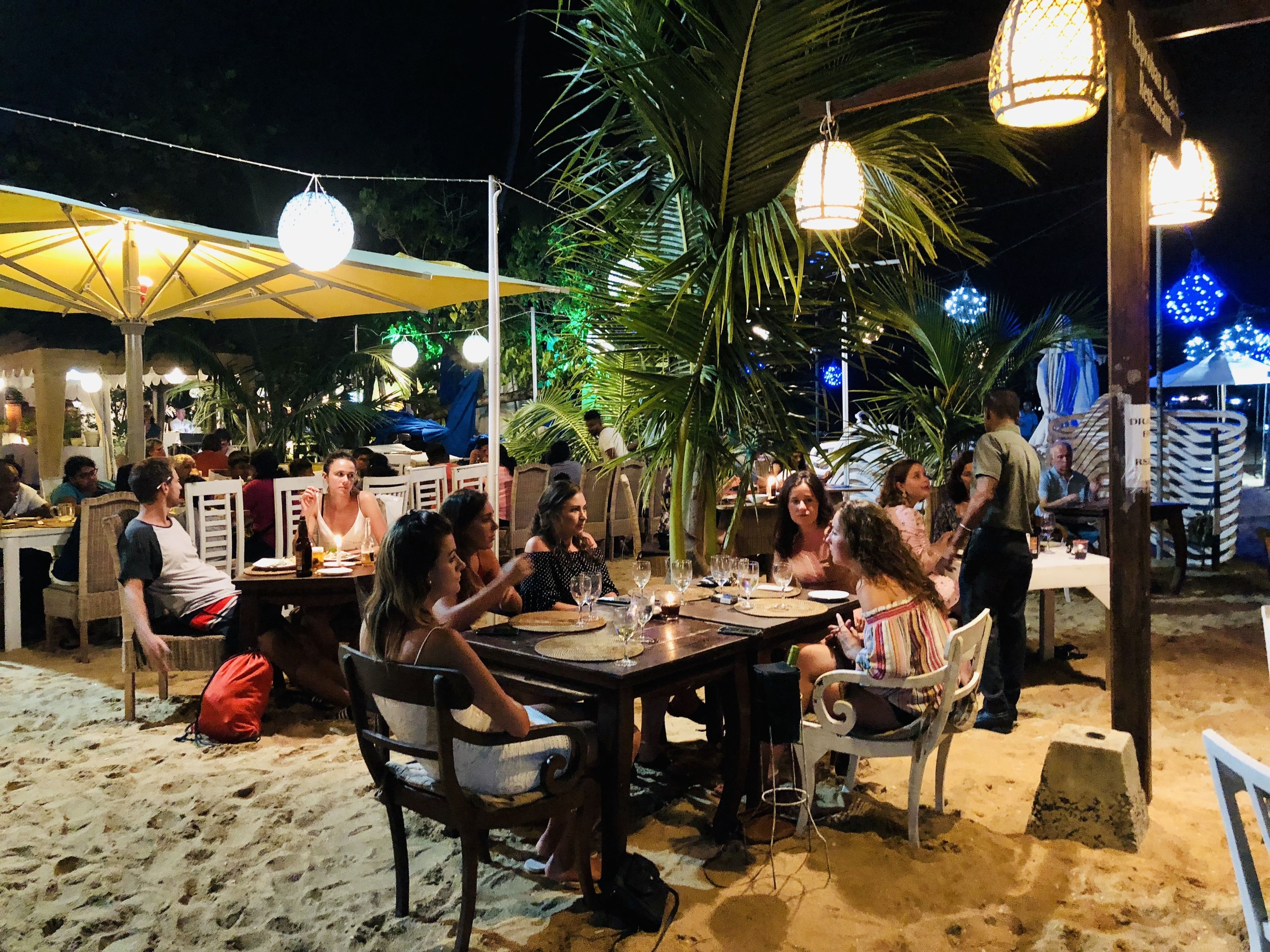 An outdoor dining area with tables and chairs, surrounded by palm trees and umbrellas.