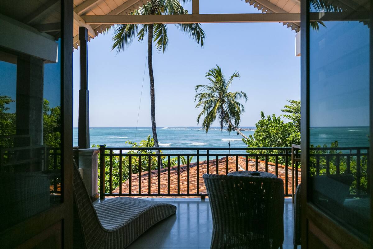 Varanda com móveis de vime com vista para uma praia com palmeiras e o oceano.