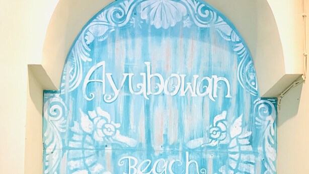 Une enseigne décorative portant l’inscription « Ayubowan Beach House » et un motif stylisé représentant une créature marine.