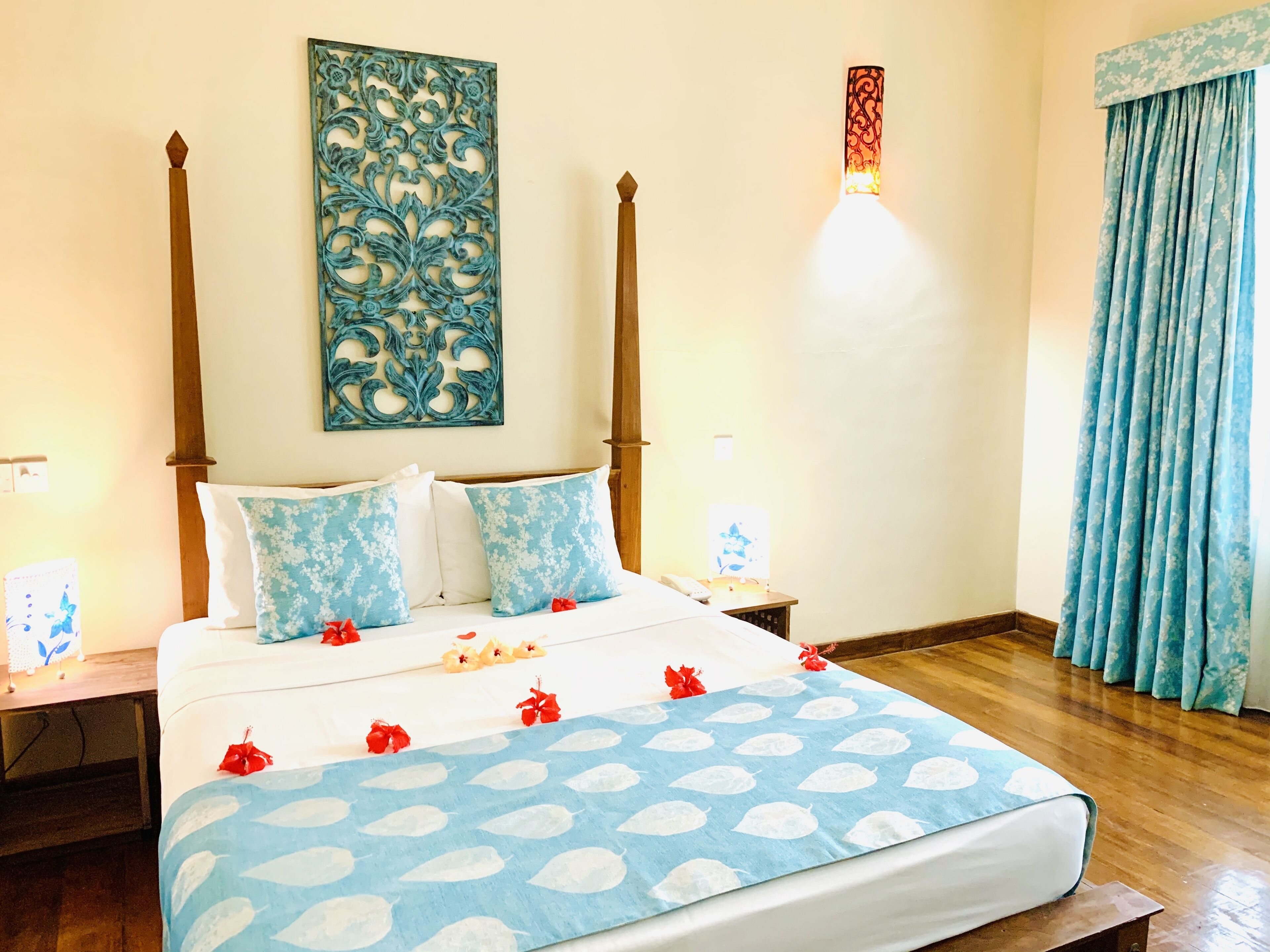 A bedroom with a four-poster bed, floral-patterned pillows, a blue leaf-patterned bedspread, and a decorative wall piece.