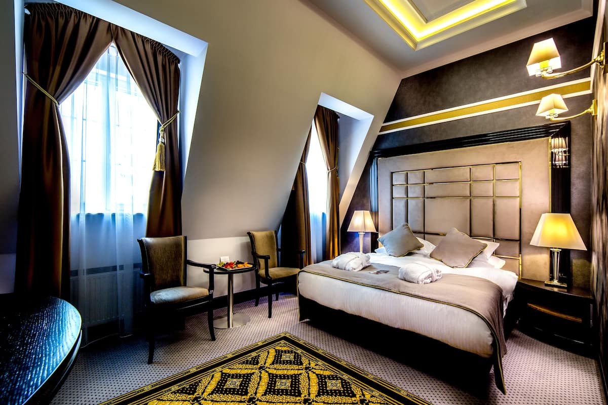 A hotel room with a large bed, two chairs, a small table with fruit, and a decorative rug.