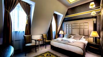 A hotel room with a large bed, two chairs, a small table with fruit, and a decorative rug.
