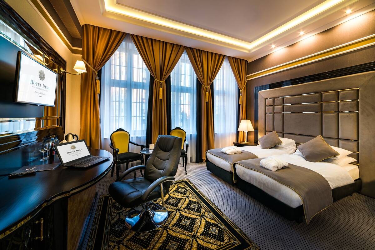 A hotel room with a desk, laptop, chair, bed, and a view of the outside through large windows.