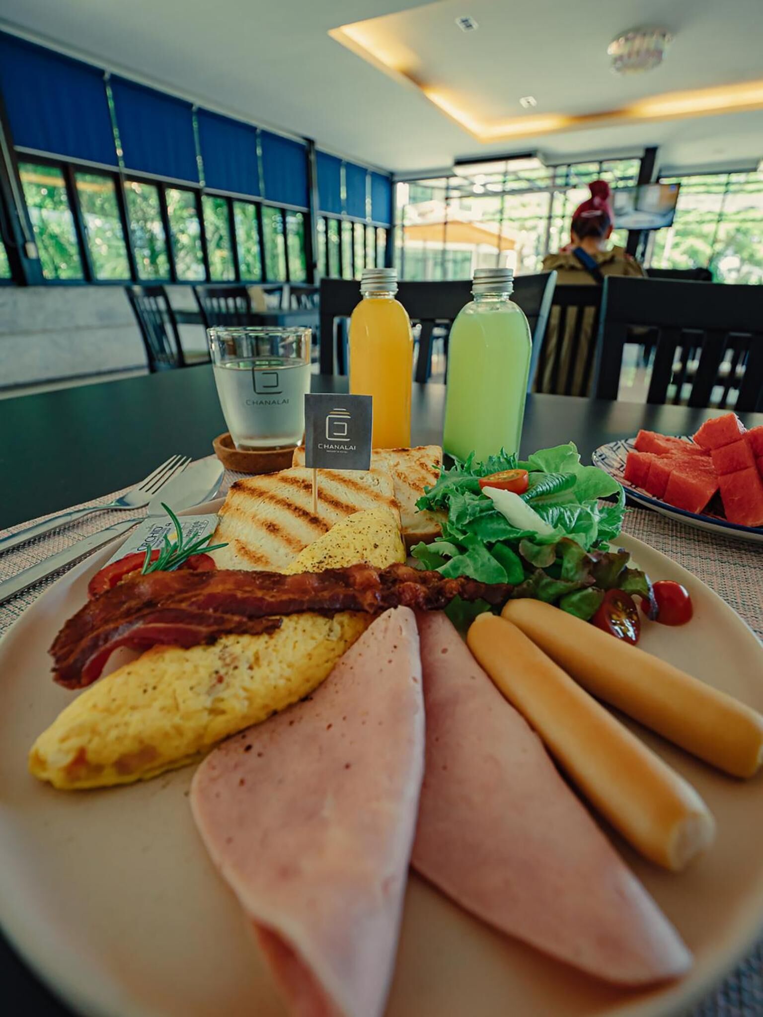 Une assiette de petit-déjeuner comprenant des œufs, du bacon, du jambon et des saucisses, accompagnée d’une salade et de pastèque, sur une table dans un restaurant offrant une vue sur la verdure.