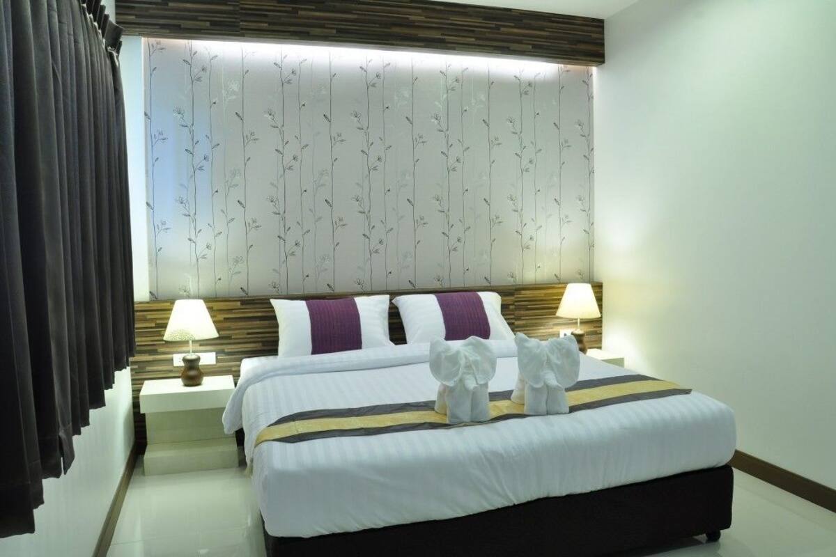 Ein Hotelzimmer mit einem großen Bett, zwei Nachttischlampen, einem Fenster mit floralem Vorhang und einem Kopfteil mit Holzpaneelen.