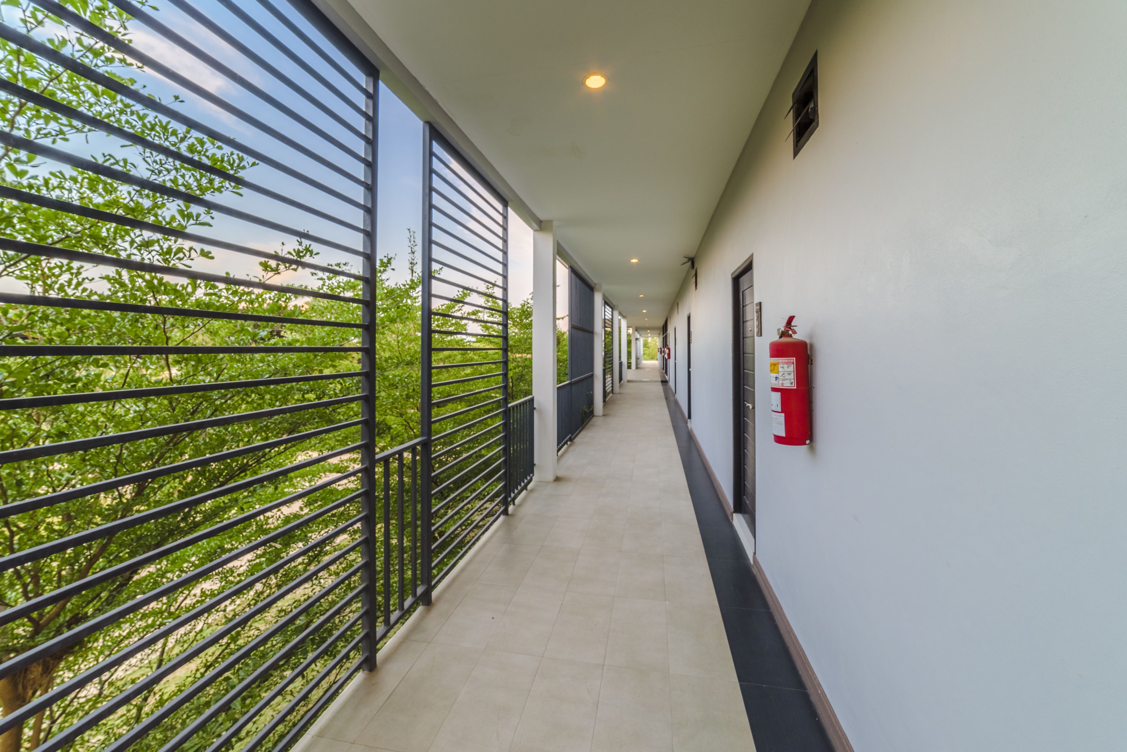 A corridor with a metal railing, a fire extinguisher, and a view of greenery outside.