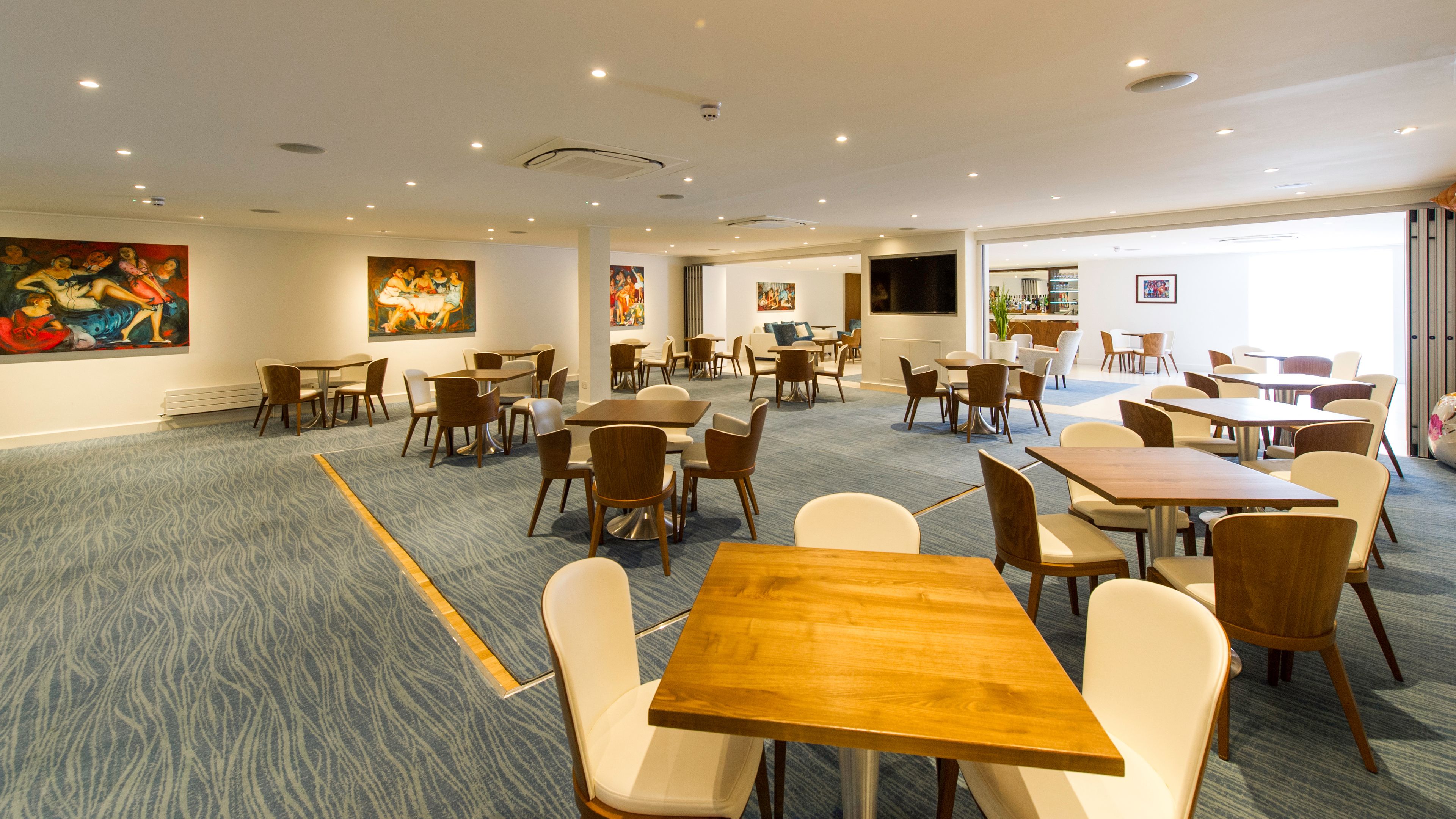 A spacious dining area with multiple tables and chairs, a patterned carpet, and several paintings on the walls.