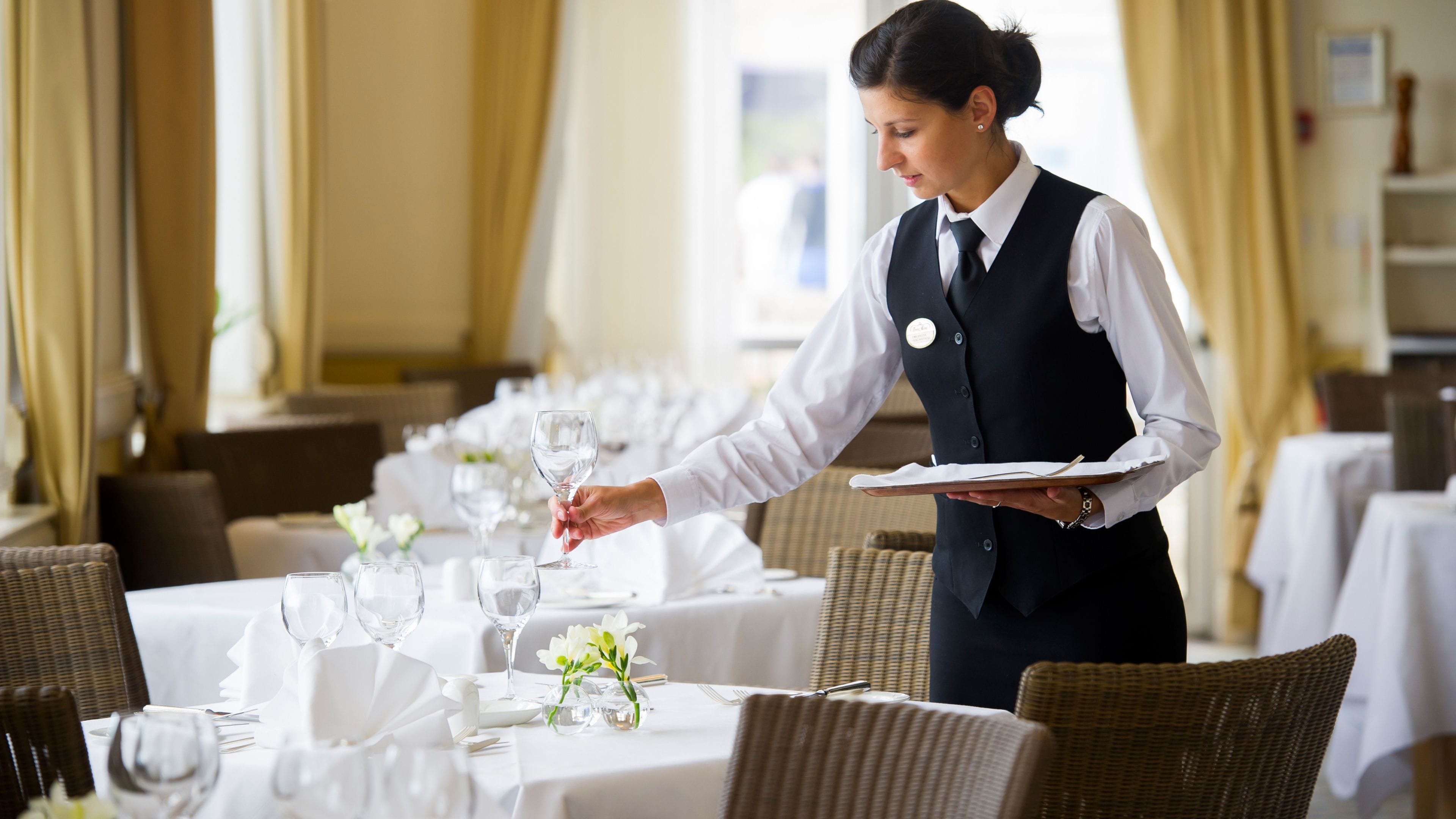 A waiter serving a table in a well-lit dining area with white tablecloths and wicker chairs.