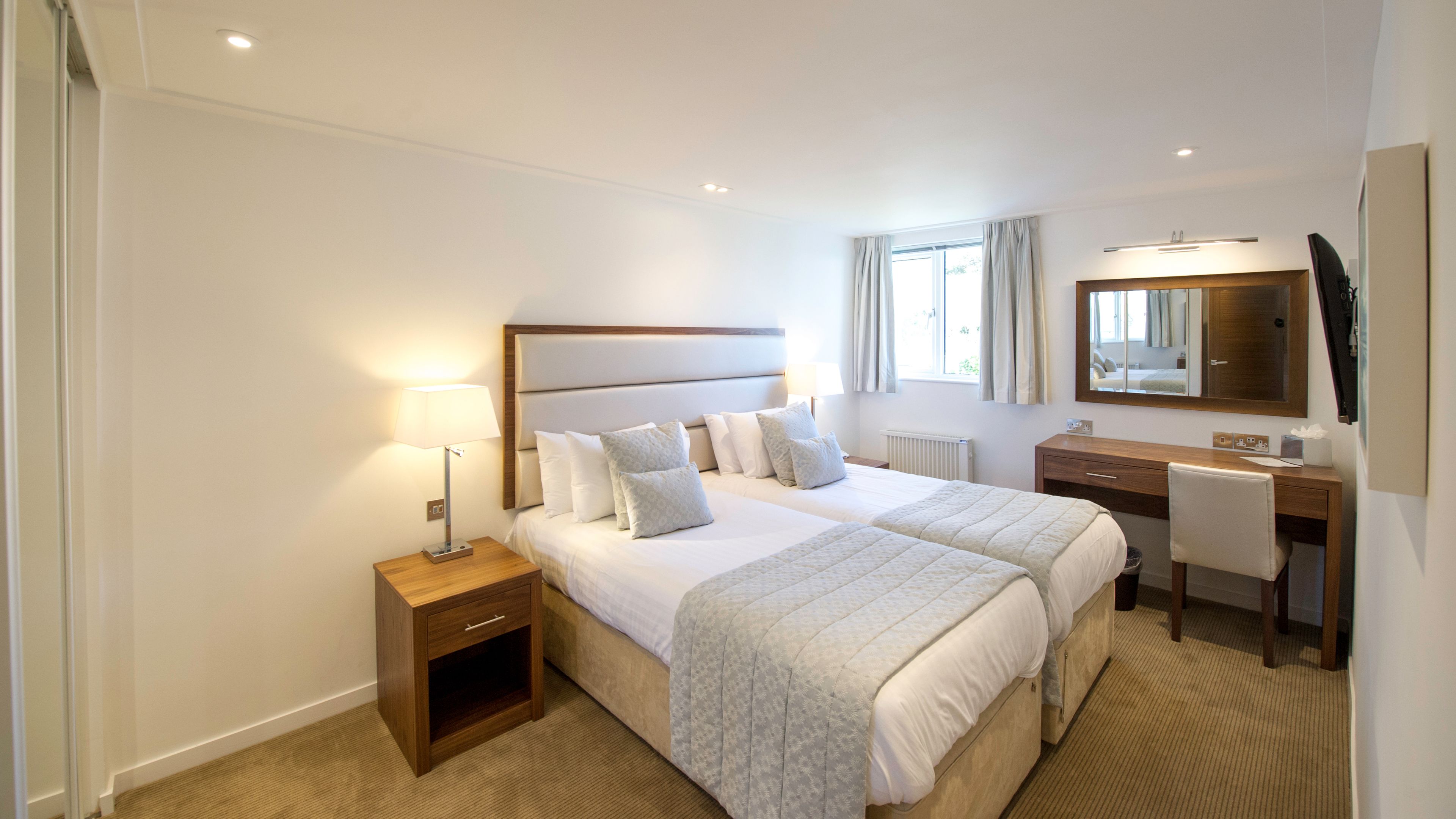 A hotel room with two beds, a desk, a chair, a lamp, a mirror, and a window.