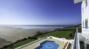 Uma piscina com padrão hexagonal, vista para a praia e um edifício branco com varanda.