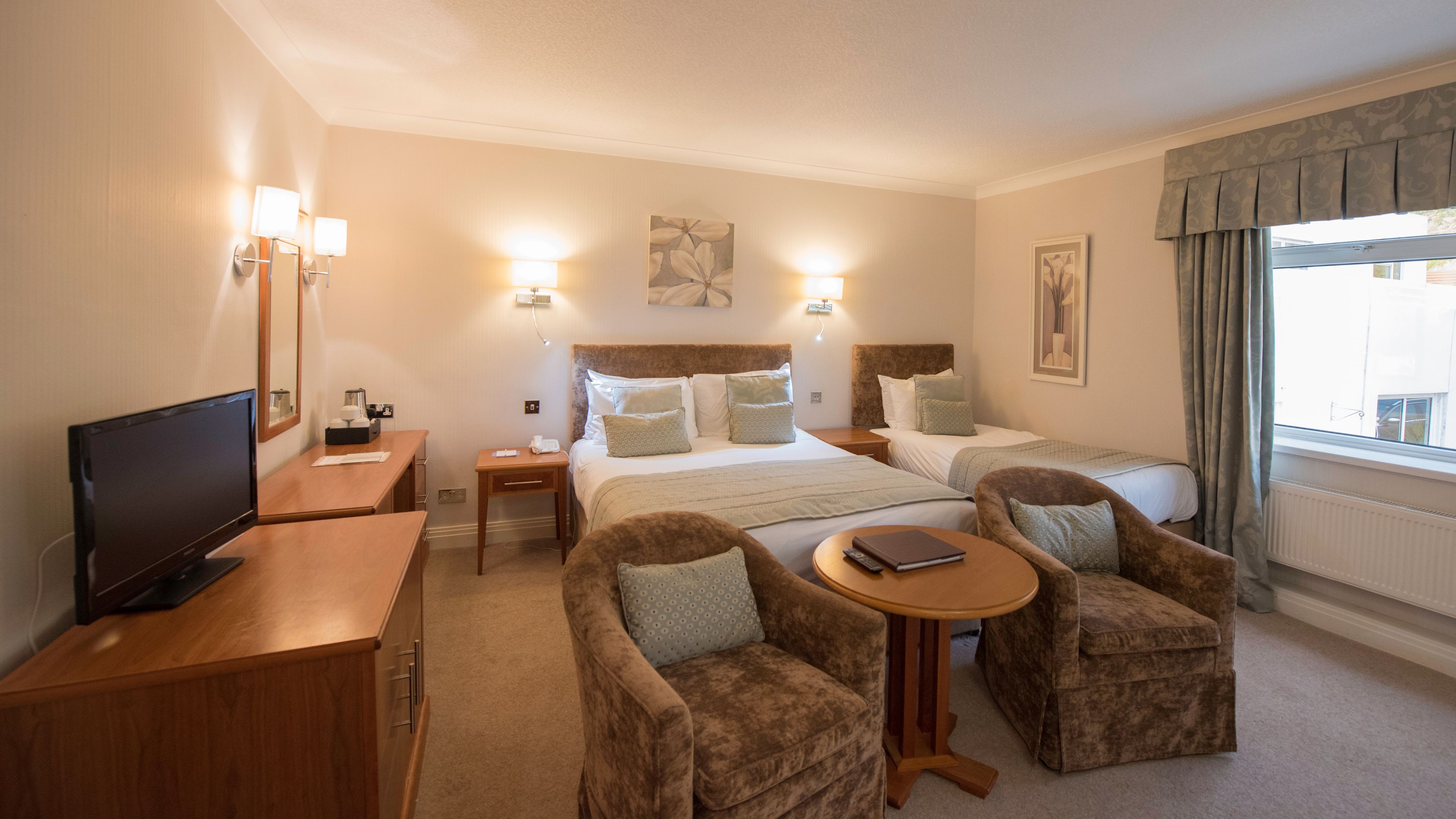 A hotel room with a bed, two bedside tables, a TV, two armchairs, a small table, and a window with curtains.