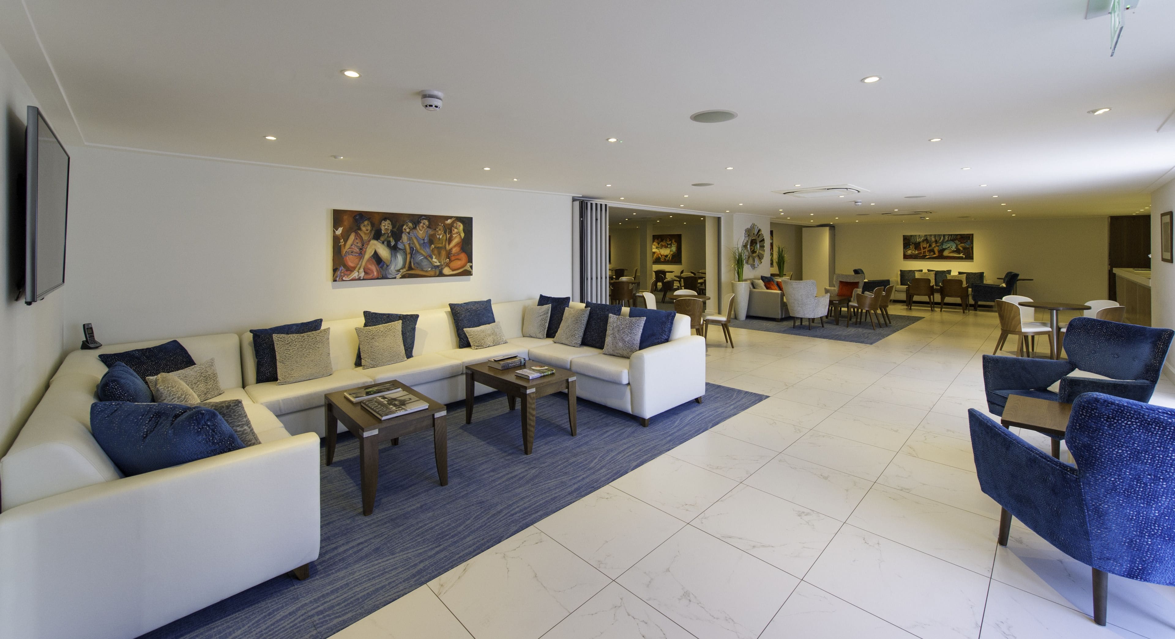 A spacious living room with a white sofa set, blue chairs, and a coffee table. There are two paintings on the walls and a dining area in the background.