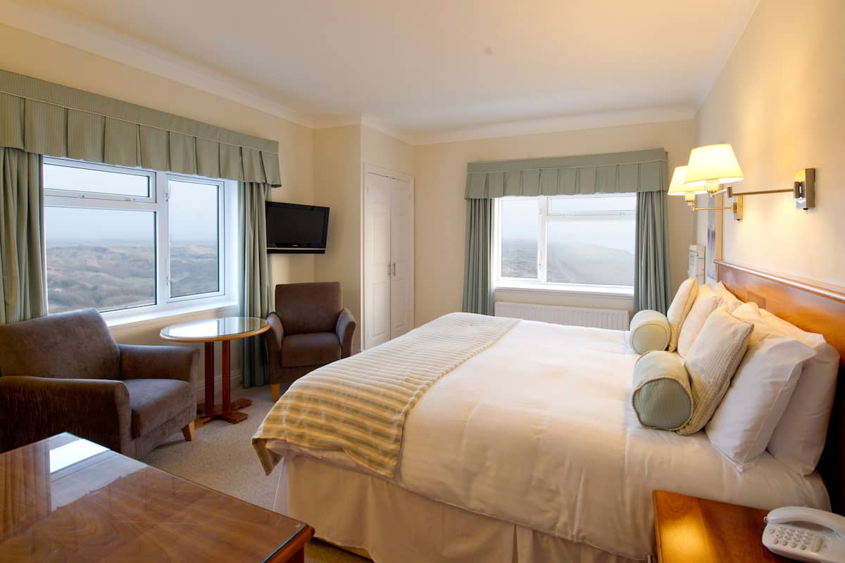 Ein Hotelzimmer mit einem großen Bett, einem Schreibtisch, einem Sessel, einem Fernseher und Blick auf die Landschaft.