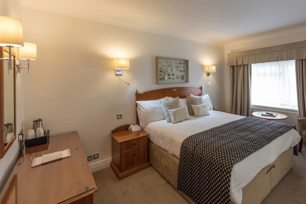 Ein Hotelzimmer mit einem großen Bett, Nachttischen, einem Schreibtisch, einem Stuhl, einem Fenster mit Vorhängen und Wandlampen.