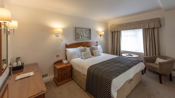 Quarto de hotel com uma cama grande, mesas de cabeceira, escrivaninha, cadeira, janela com cortinas e luminárias fixadas na parede.