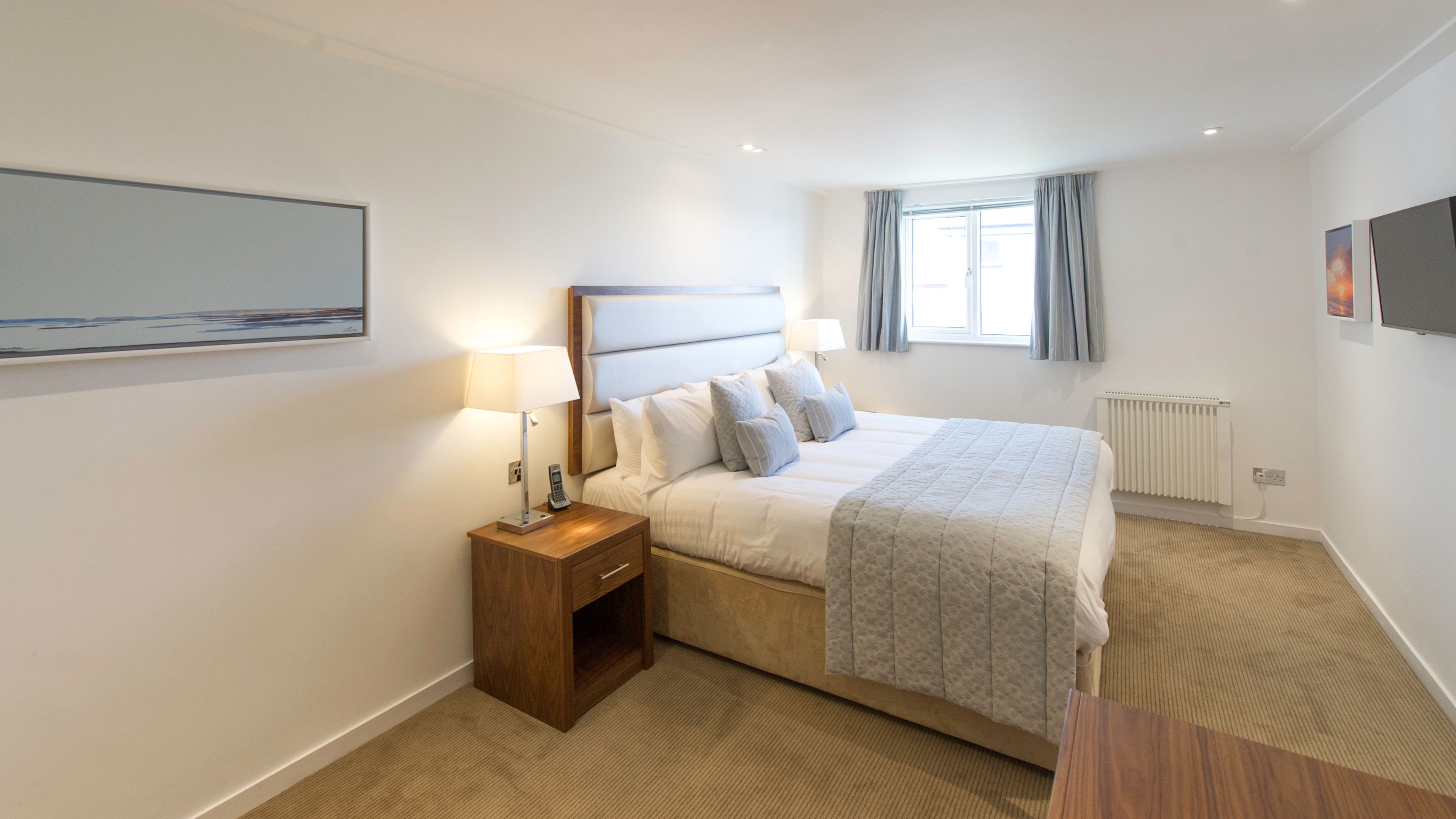 A hotel room with a bed, bedside lamps, a nightstand, a window with curtains, a radiator, and a painting on the wall.