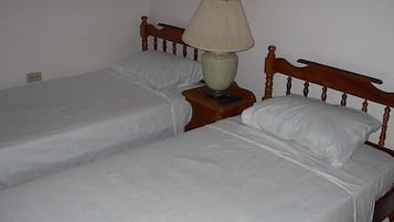 Una habitación con dos camas individuales, cada una con cabecero de madera y ropa de cama blanca. Hay una mesita de noche con una lámpara entre las camas.