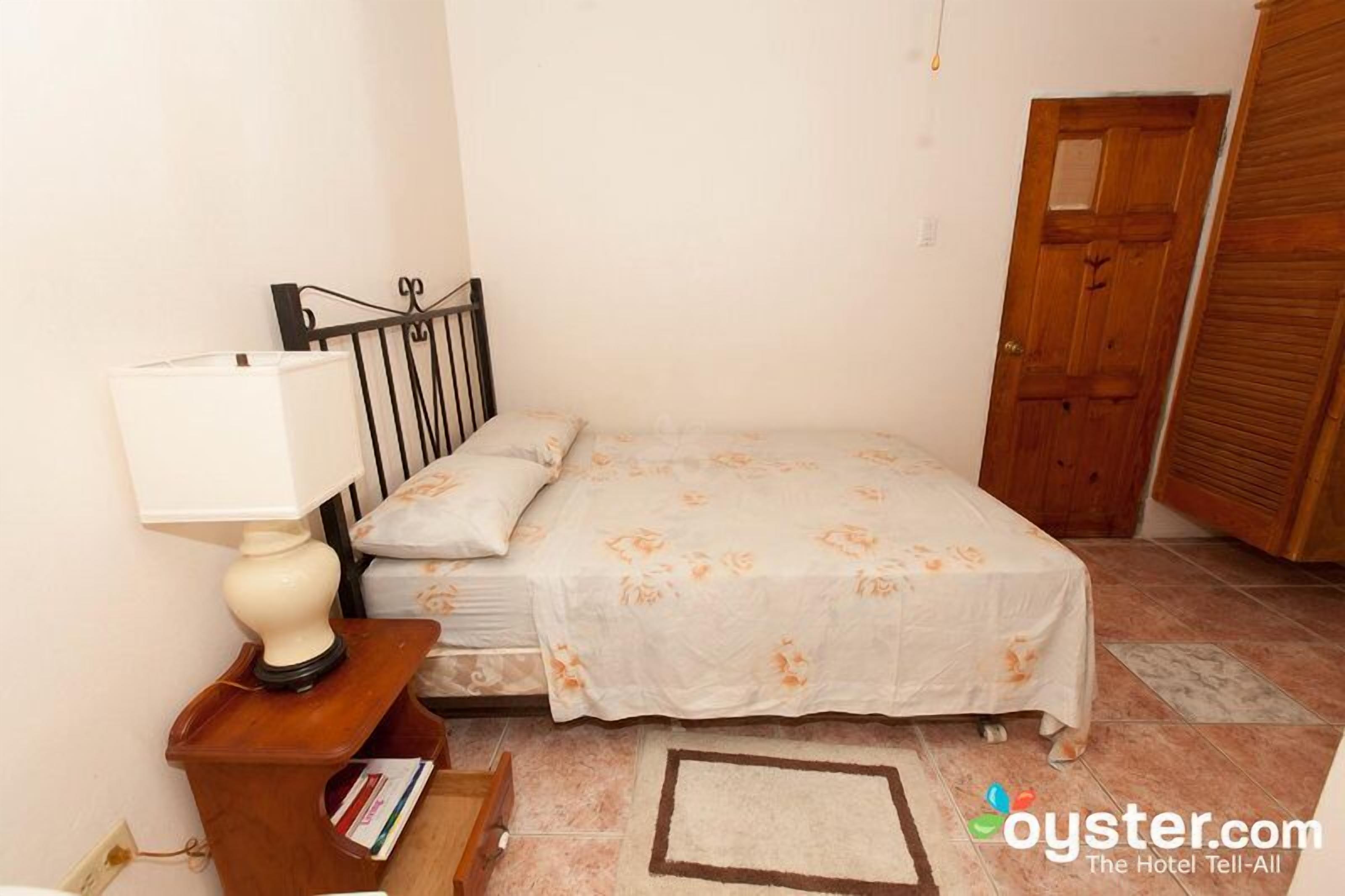 A bedroom with a bed, a nightstand with a lamp, a wooden door, and a tiled floor.