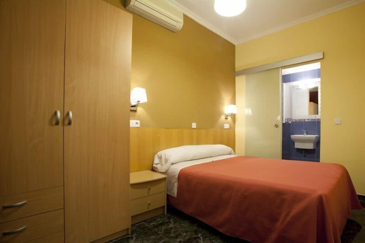 Quarto de hotel com cama, um guarda-roupa, mesa de cabeceira e banheiro visível através de uma porta aberta.