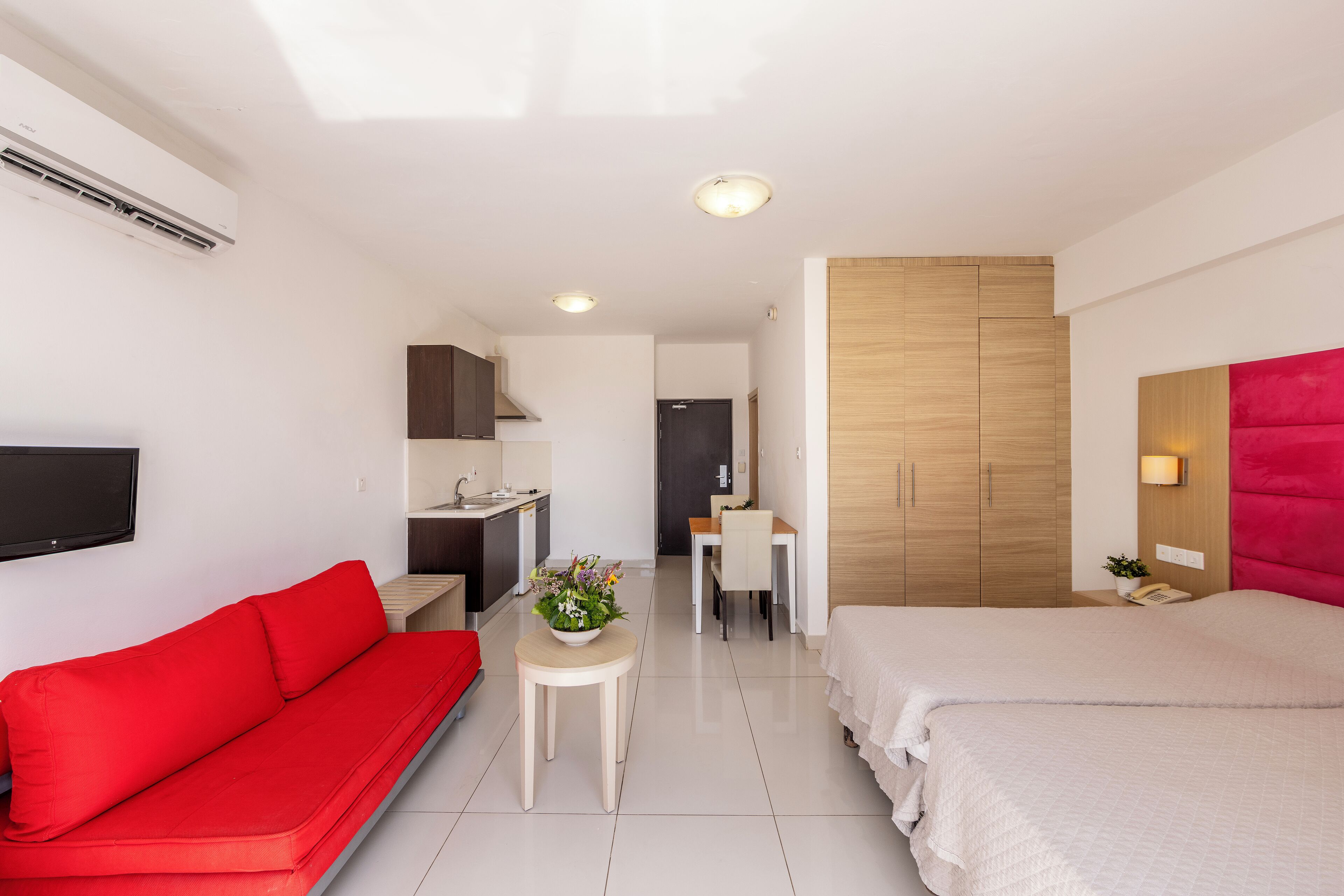 A modern hotel room with a red sofa, a bed, a small table with a flower vase, a desk with a chair, and a wardrobe.