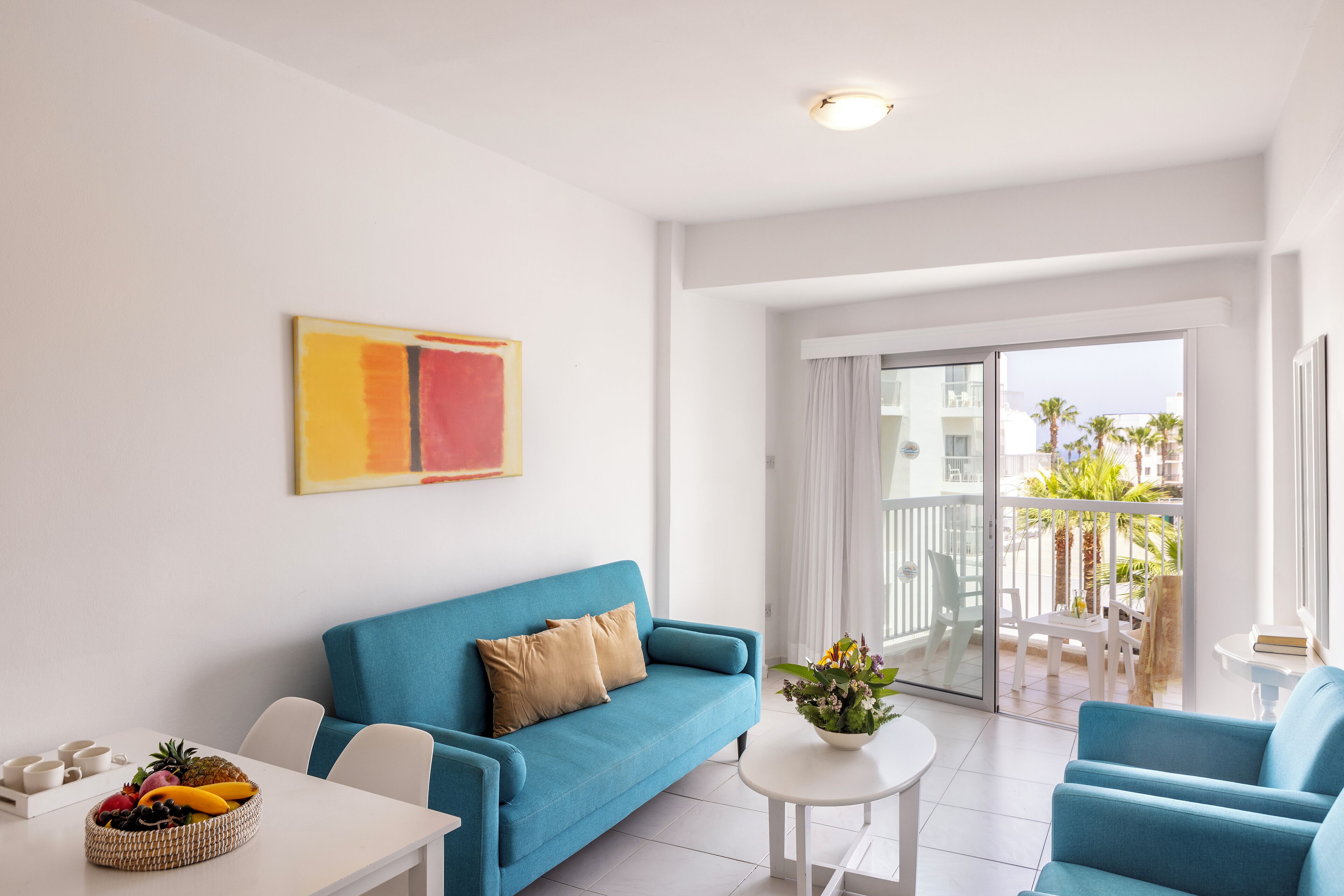 A modern living room with a blue sofa, a white table with fruit, and a balcony with a view of palm trees.